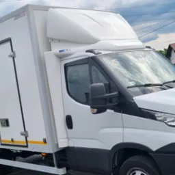 Biały samochód dostawczy Iveco z zabudową kontenerową, zaparkowany na kostce brukowej, z widoczną tabliczką MRauto na zabudowie i fragmentem zabudowań w tle.