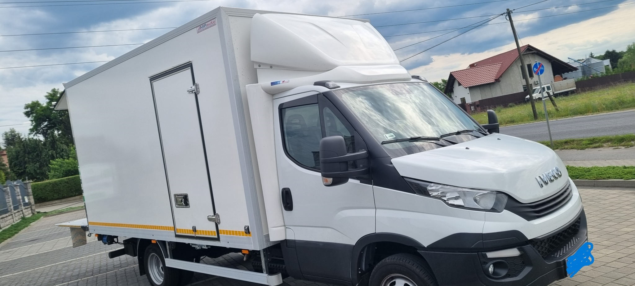 Biały samochód dostawczy Iveco z zabudową kontenerową, zaparkowany na kostce brukowej, z widoczną tabliczką MRauto na zabudowie i fragmentem zabudowań w tle.