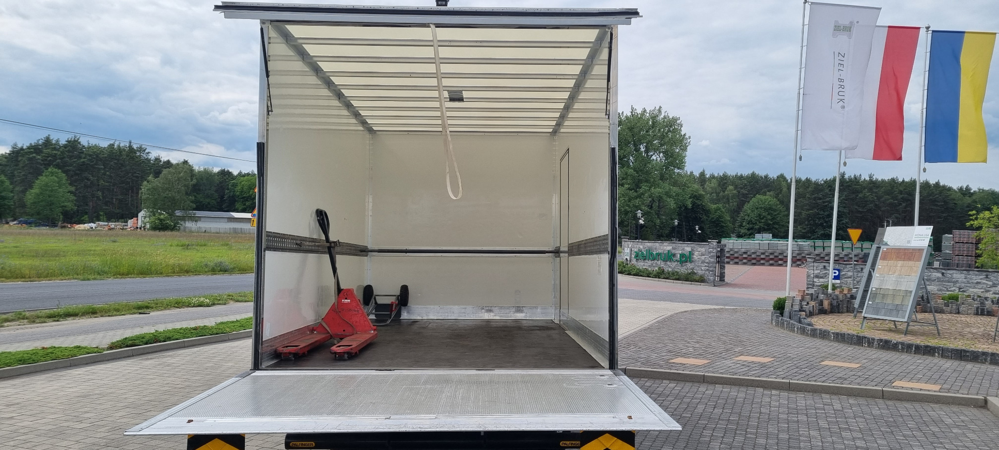 Wnętrze pustej, otwartej paki busa transportowego z wózkiem paletowym, widok z zewnątrz na otoczenie z zielenią i flagami.