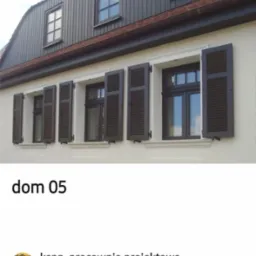 Elewacja domu z oknami i brązowymi okiennicami. Część okiennic jest otwarta. Widoczna drewniana obudowa poddasza w kolorze ciemnobrązowym.