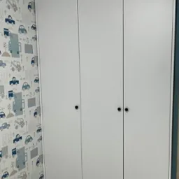 Trzydrzwiowa, biała szafa z okrągłymi uchwytami, na tle tapety w dziecięce motywy samochodowe. Ujęcie z dołu, ukazujące całą wysokość mebla.