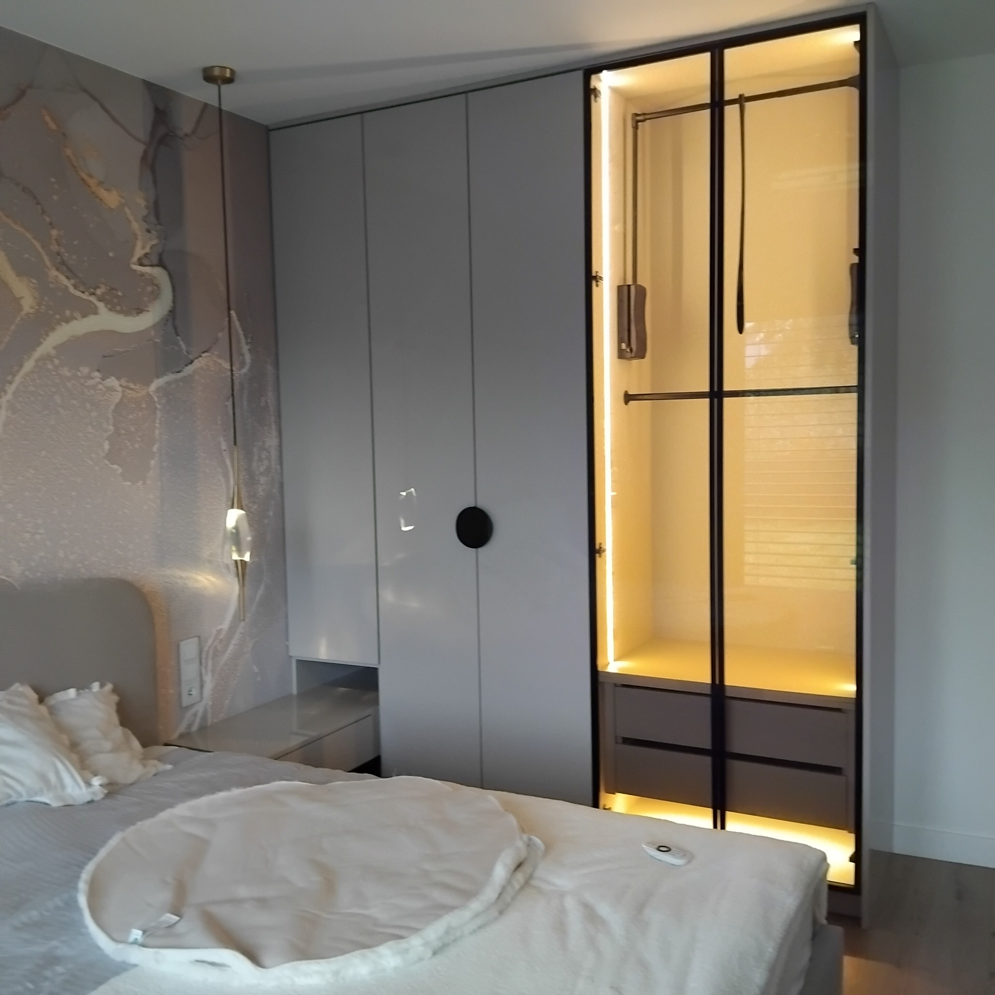 Szafa wnękowa z oświetleniem LED, szare fronty i czarne detale, widoczna z perspektywy łóżka w sypialni z dekoracyjną lampą wiszącą i tapetą.