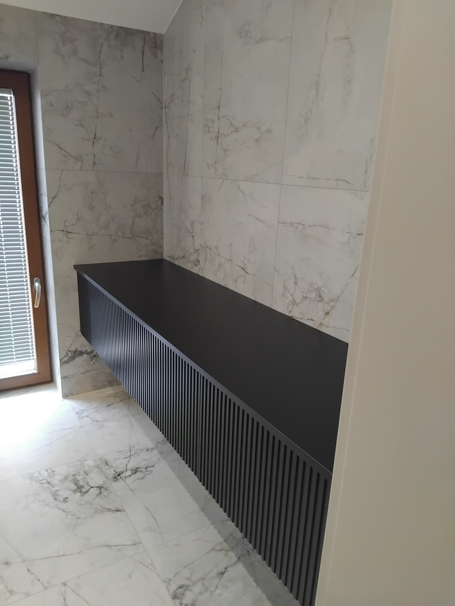 Czarna, podwieszana szafka łazienkowa z ryflowanym frontem na tle marmurowych płytek i okna z żaluzjami, minimalistyczny design.