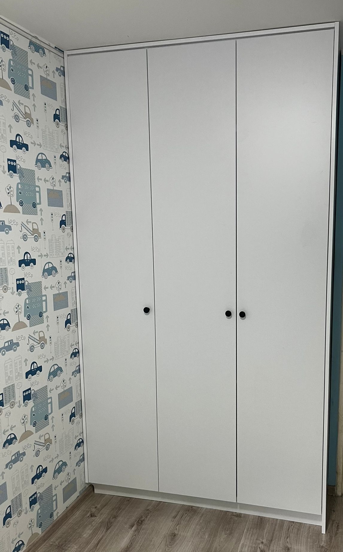 Trzydrzwiowa, biała szafa z okrągłymi uchwytami, na tle tapety w dziecięce motywy samochodowe. Ujęcie z dołu, ukazujące całą wysokość mebla.