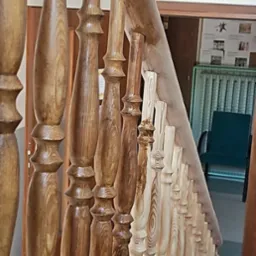 Drewniane balustrady schodowe, zróżnicowane wybarwienie bejcą, od jasnego do ciemnego brązu, widoczne słoje drewna, w tle drzwi i tabliczka informacyjna.