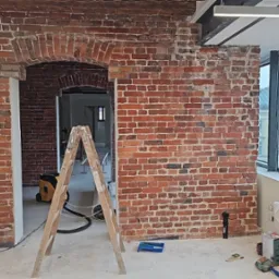 Ceglana ściana z łukowym przejściem w trakcie renowacji, widoczne rusztowanie i narzędzia. Fragment okna z widokiem na miasto w tle.