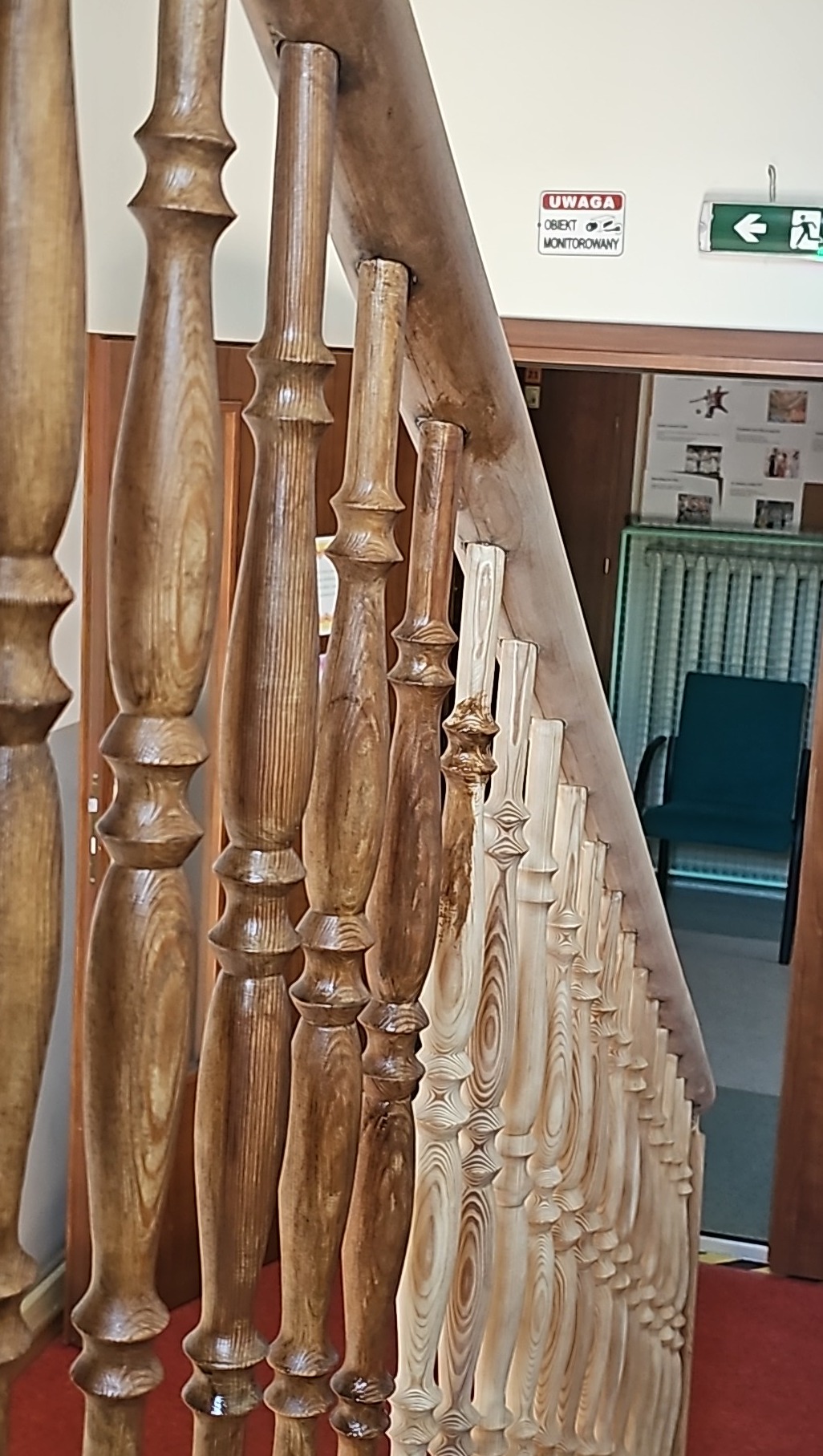 Drewniane balustrady schodowe, zróżnicowane wybarwienie bejcą, od jasnego do ciemnego brązu, widoczne słoje drewna, w tle drzwi i tabliczka informacyjna.
