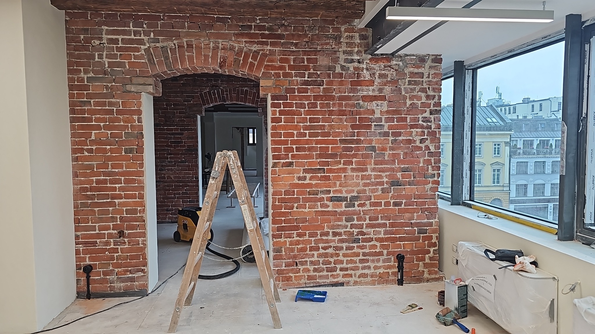 Ceglana ściana z łukowym przejściem w trakcie renowacji, widoczne rusztowanie i narzędzia. Fragment okna z widokiem na miasto w tle.