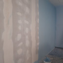 Piotr Fiebig - Ściana po szpachlowaniu z widocznymi łatami, częściowo pomalowana na niebiesko. Wiadro z farbą i wałek malarski na podłodze. Prace wykończeniowe w pomieszczeniu.