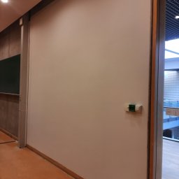 Piotr Fiebig - Świeżo pomalowana, gładka ściana w jasnym odcieniu beżu, z widocznym panelem sterowania i fragmentem przeszklonego wejścia, sugerująca wykończenie wnętrza.