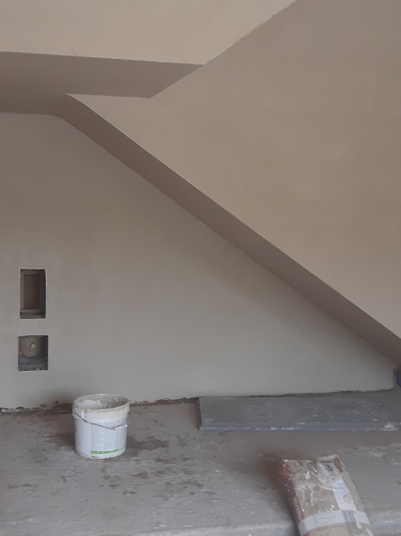 Ściana po szpachlowaniu z widocznymi otworami na instalacje i wiadrem z masą szpachlową. Ukośna linia sufitu. Neutralna kolorystyka.