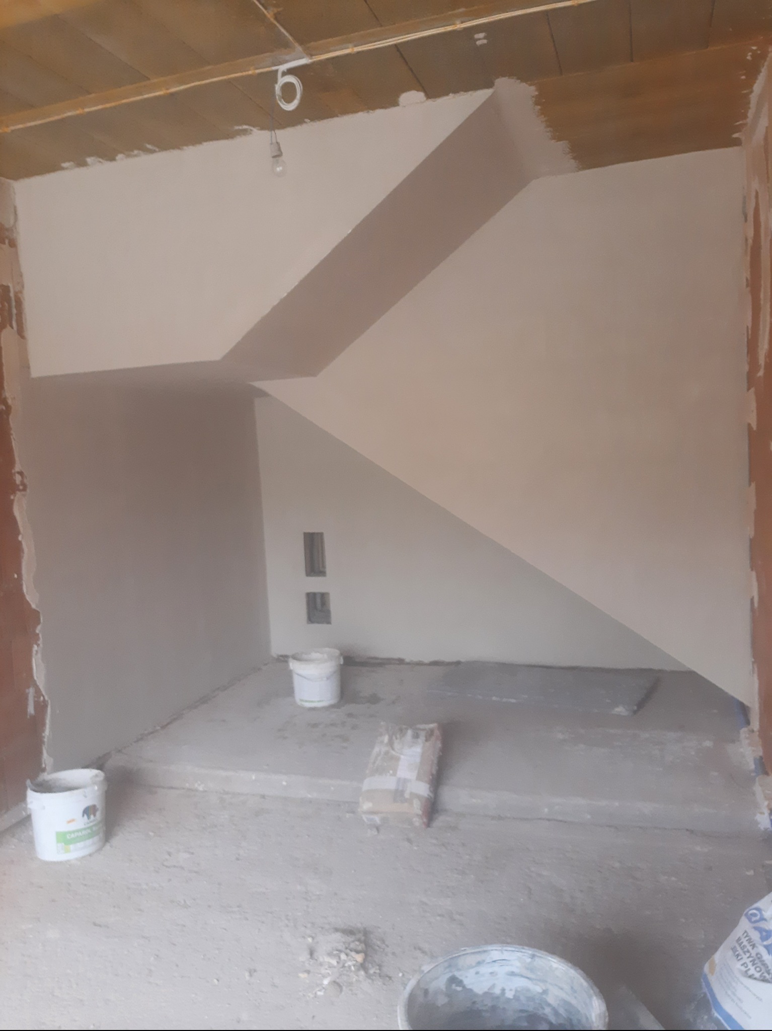 Wnętrze w trakcie prac wykończeniowych: ściany pokryte gładzią, widoczne wiadra z zaprawą i worki z materiałami budowlanymi. Surowy stan, przygotowanie do malowania.