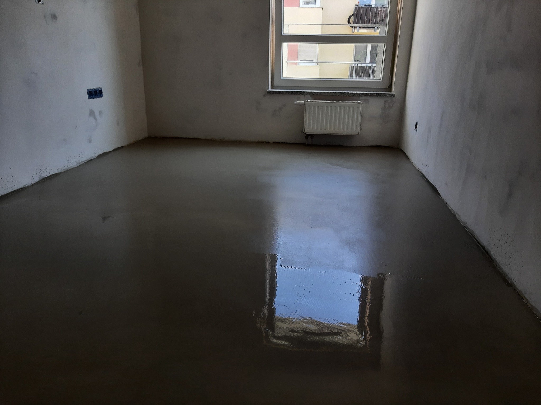 Wylana, mokra posadzka samopoziomująca w pokoju z oknem i grzejnikiem. Ściany przygotowane do dalszych prac wykończeniowych, widoczne puszki elektryczne.