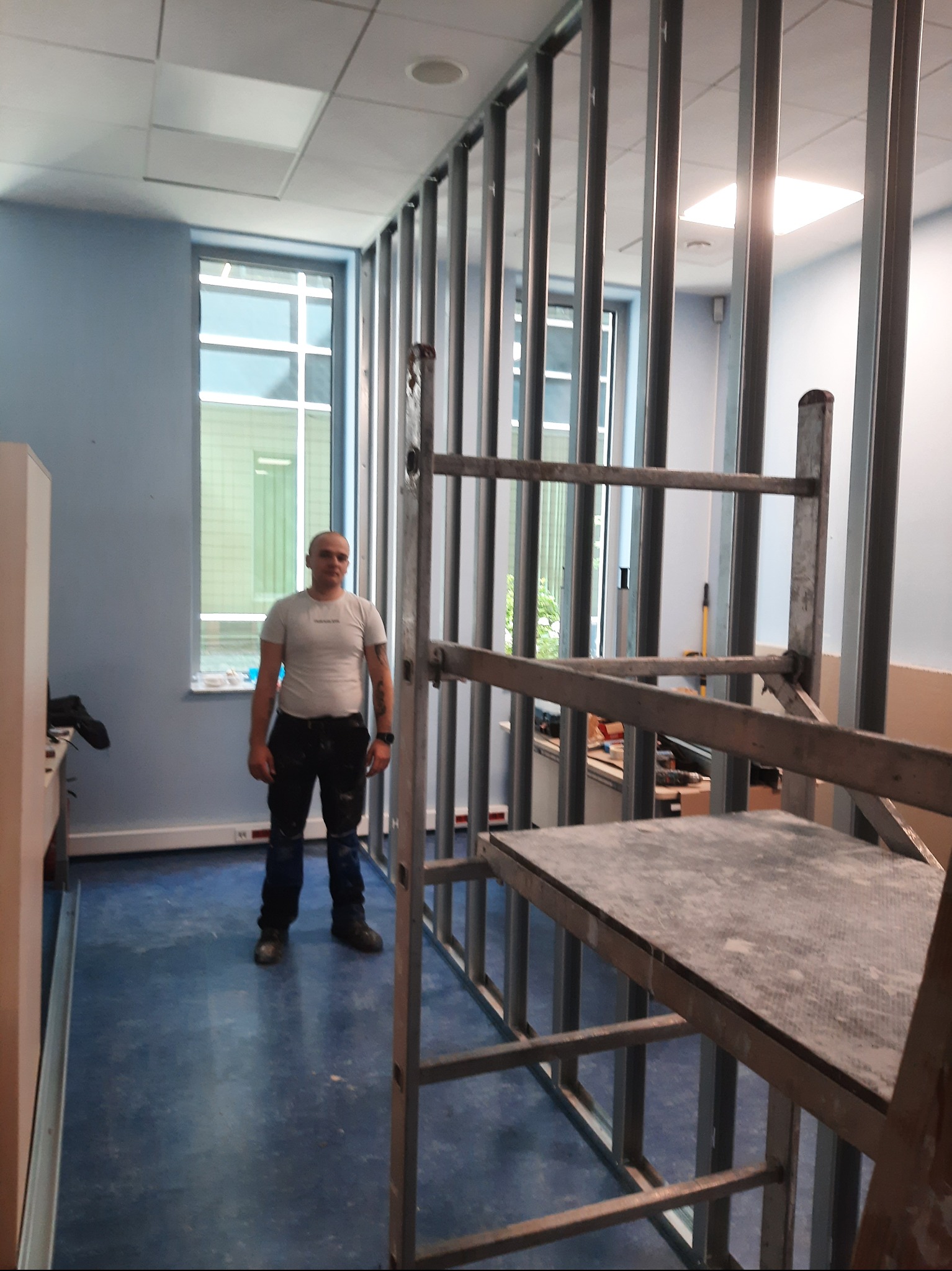 Montaż metalowej konstrukcji ścianki działowej w biurze, widoczny pracownik i drabina. Ściany pomalowane na niebiesko, duże okna z widokiem na zewnątrz.