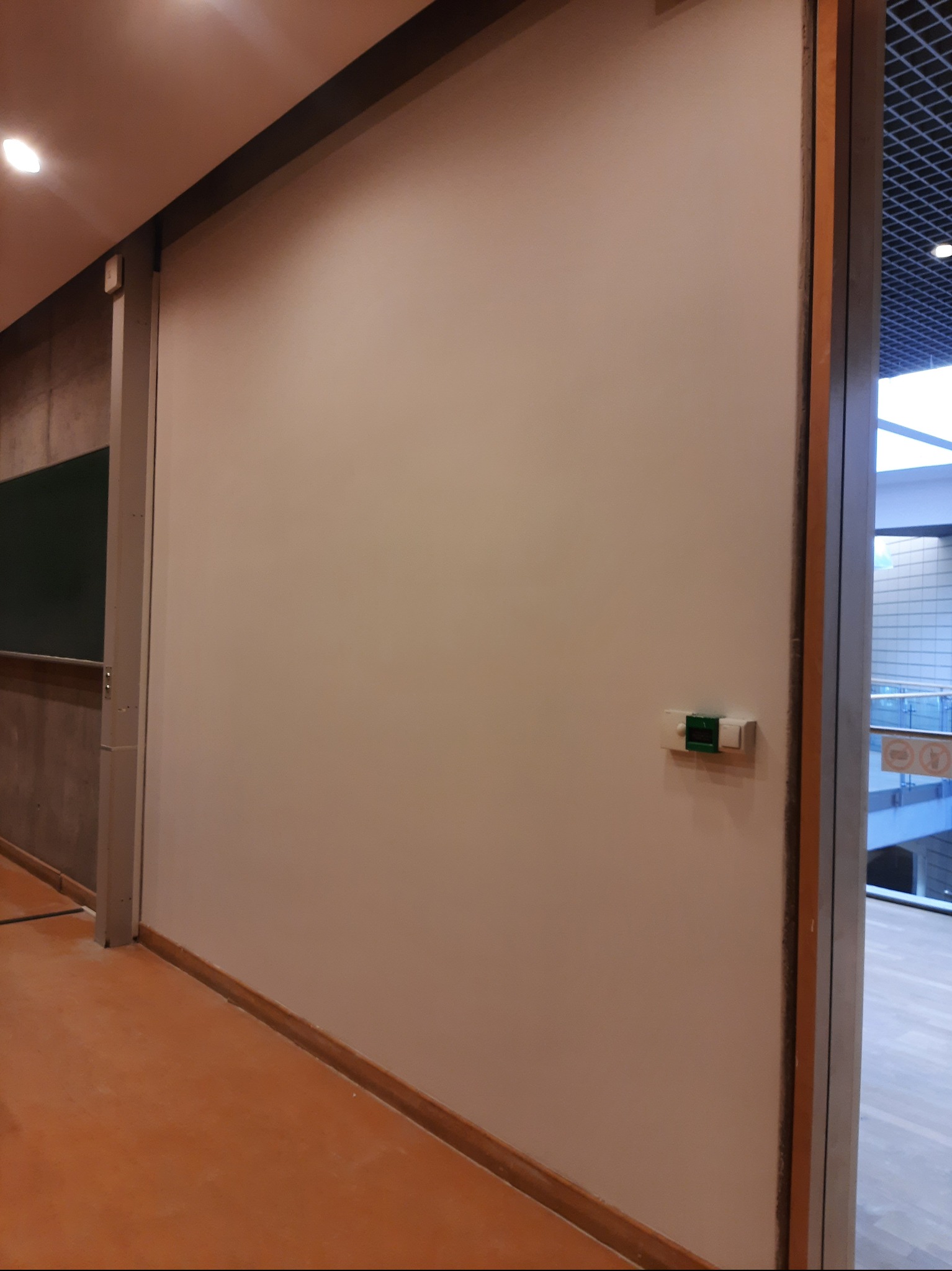 Świeżo pomalowana, gładka ściana w jasnym odcieniu beżu, z widocznym panelem sterowania i fragmentem przeszklonego wejścia, sugerująca wykończenie wnętrza.