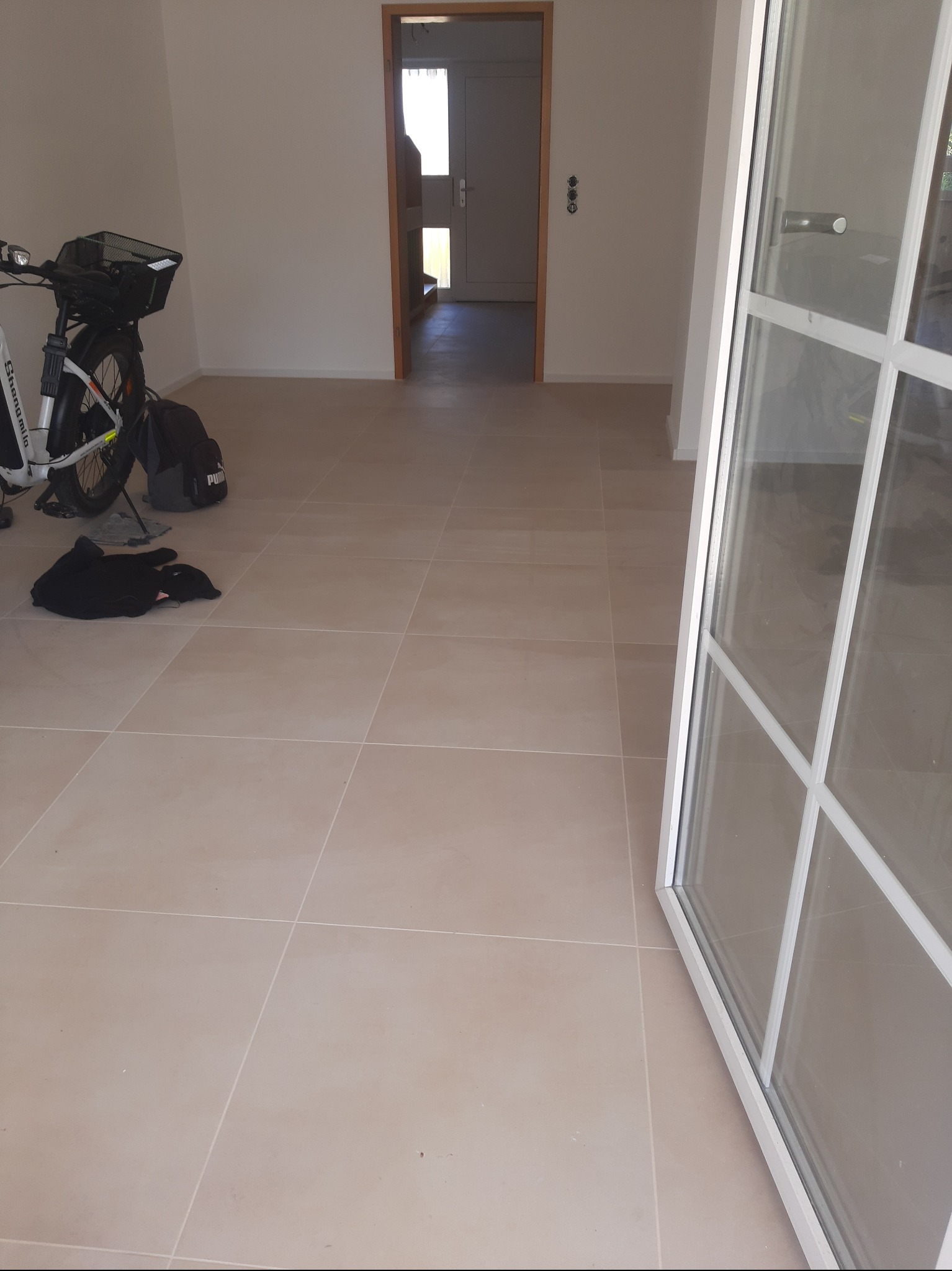 Jasne, kwadratowe płytki podłogowe w nowym wnętrzu. Widoczne drzwi, okno i rower elektryczny. Minimalistyczny styl wykończenia.