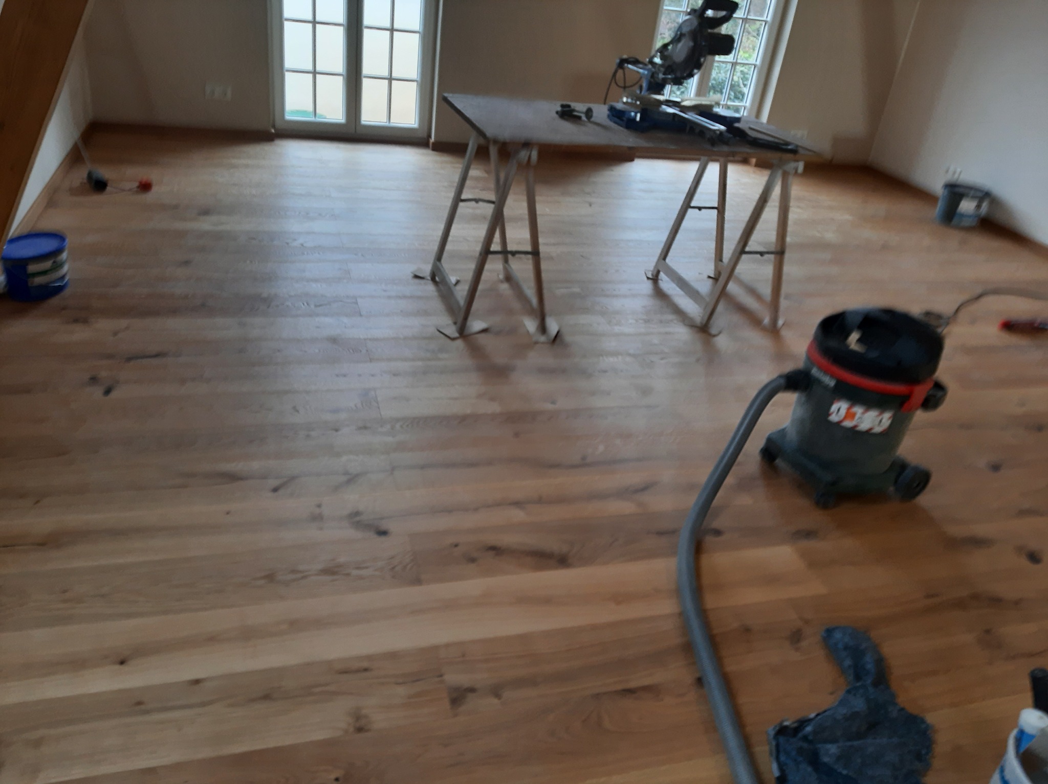 Ukończona drewniana podłoga w jasnym odcieniu, z widocznym odkurzaczem przemysłowym, stołem roboczym i narzędziami w tle, po zakończonych pracach wykończeniowych.