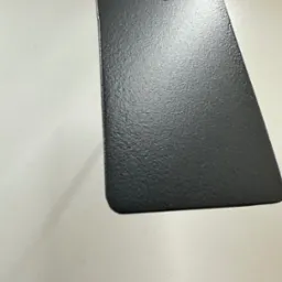 Czarny, metalowy element z otworem, zawieszony na lince, prezentujący chropowatą fakturę powierzchni po obróbce.