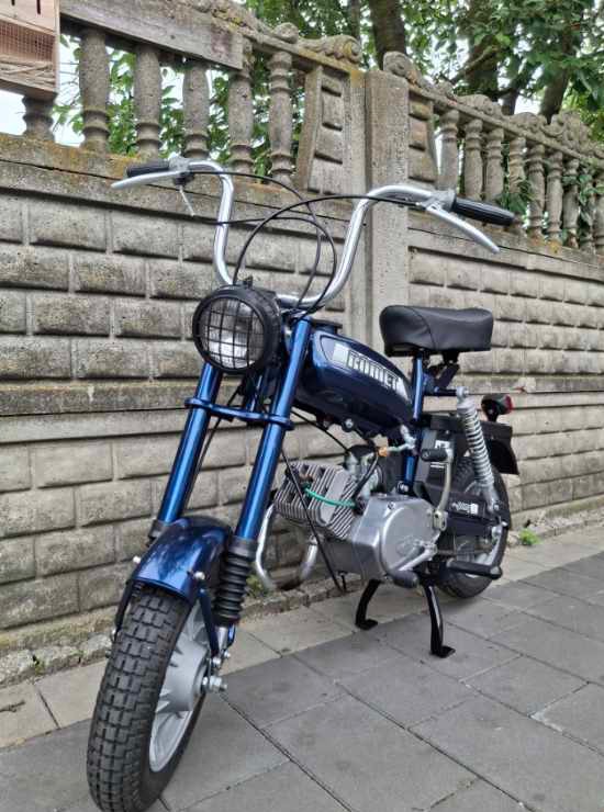 Odrestaurowany, granatowy motorower Romet z chromowanymi elementami, zaparkowany na chodniku przed murem z dekoracyjnym betonowym płotem. Widok z przodu, lekko z dołu.