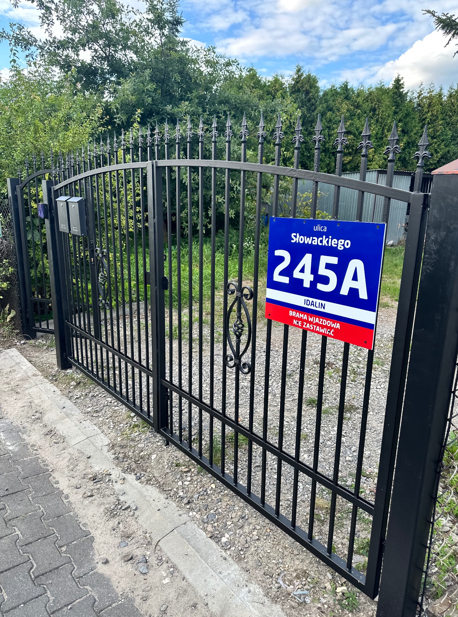 Czarna, metalowa brama z dekoracyjnymi grotami, tabliczka z adresem 'ul. Słowackiego 245A'. Brama wjazdowa do posesji, widok z zewnątrz, w tle zieleń.