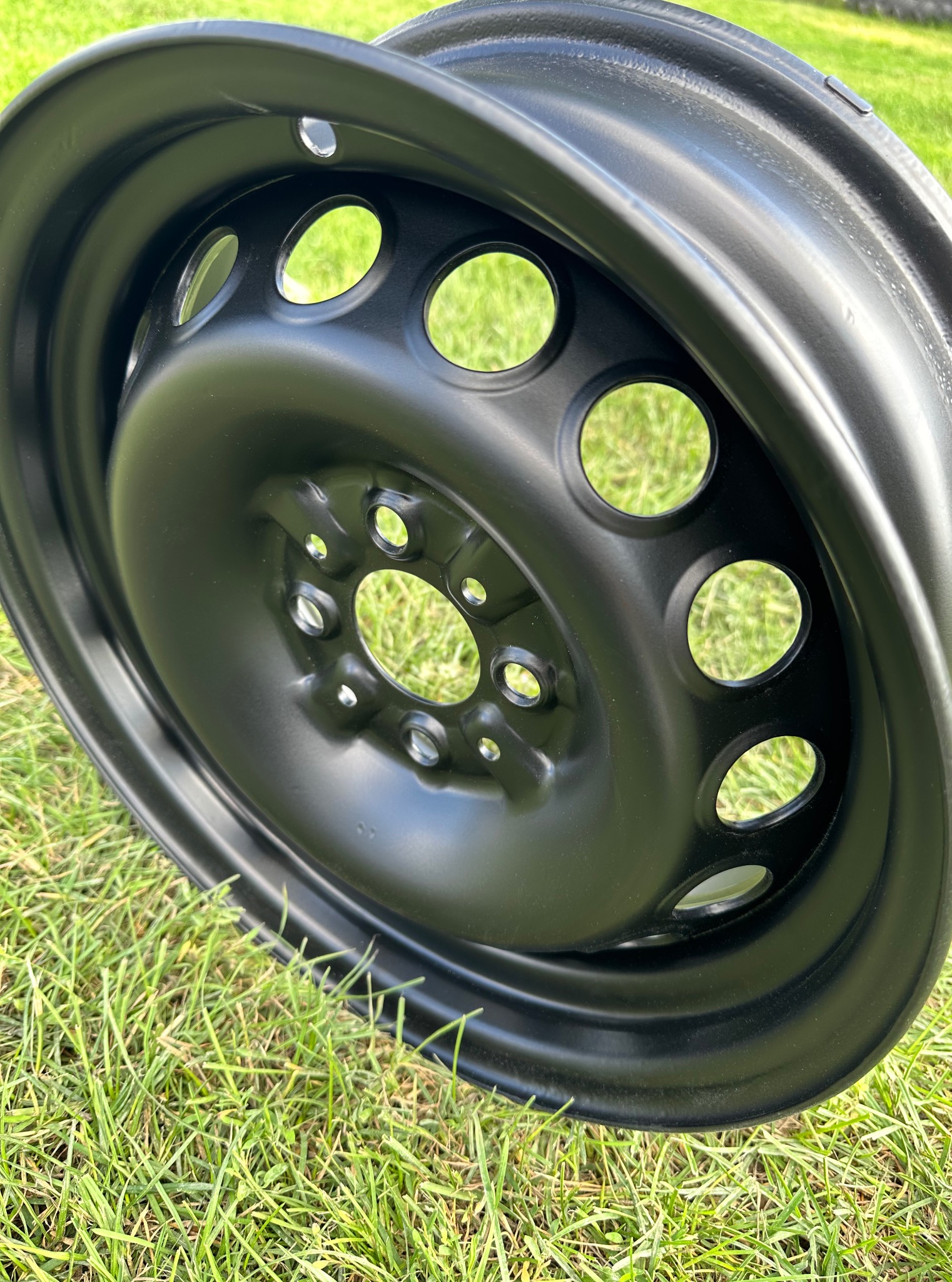 Czarna, pomalowana felga samochodowa z otworami na tle zielonej trawy, ukazująca gładką powierzchnię i równomierne pokrycie farbą.