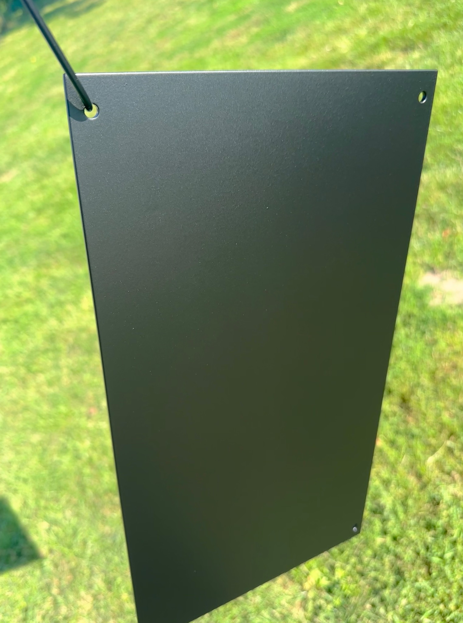 Ciemny, matowy element metalowy z otworami, zawieszony na tle zielonej trawy. Widoczne delikatne odbicia światła na powierzchni.