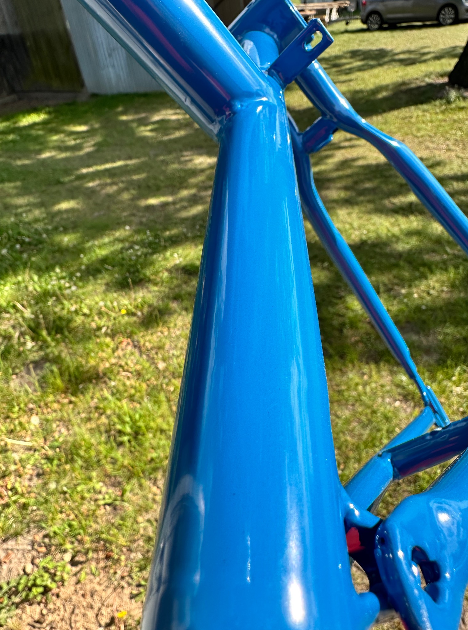 Metalowa konstrukcja pomalowana na niebiesko, widoczne spawy, gładka powierzchnia, tło z zielonej trawy i cień drzew. Detal po obróbce.