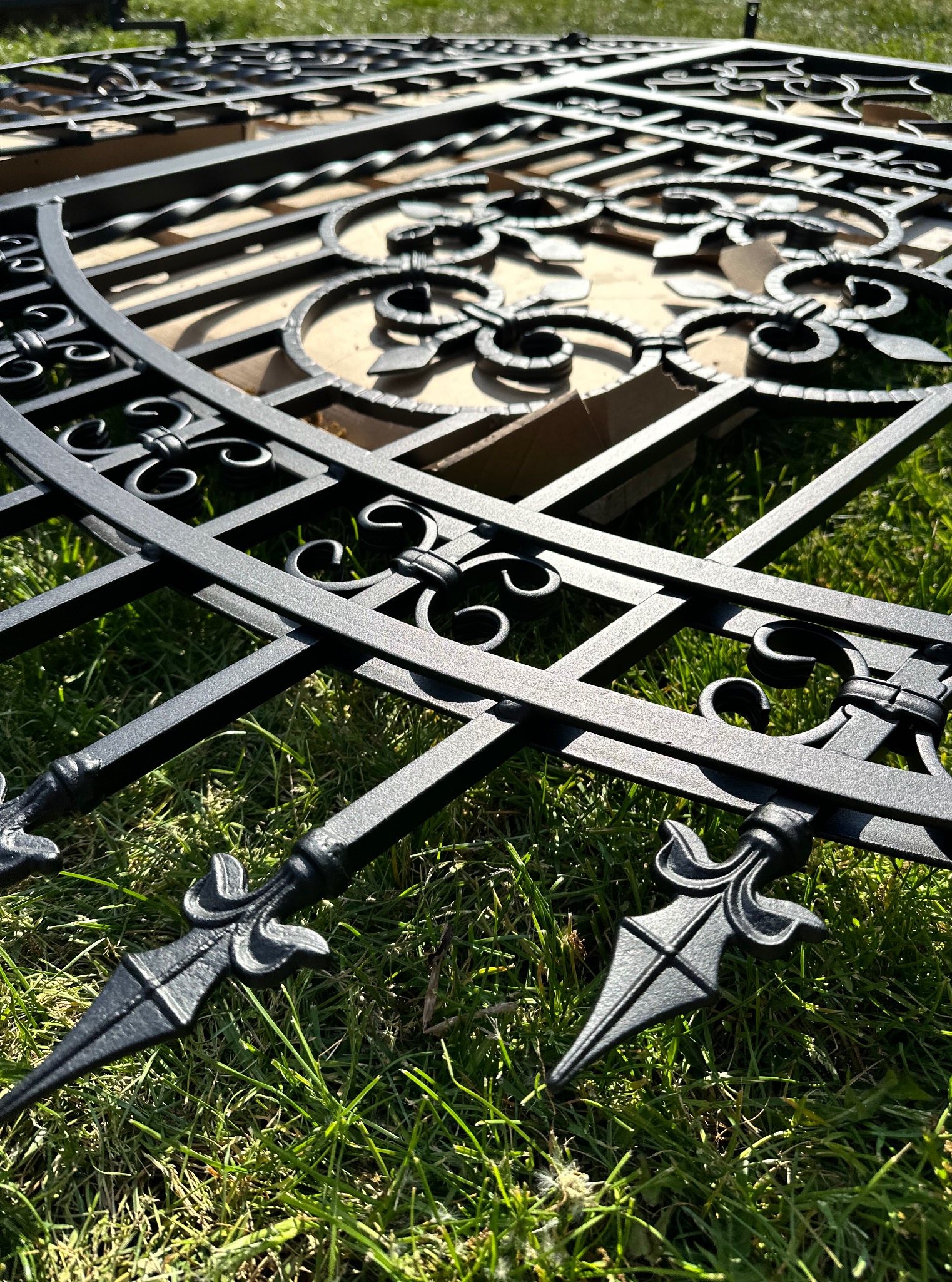 Elementy metalowe dekoracyjnego ogrodzenia, świeżo po obróbce, leżą na trawie w słoneczny dzień. Widoczne detale zdobień i matowa, czarna powierzchnia.