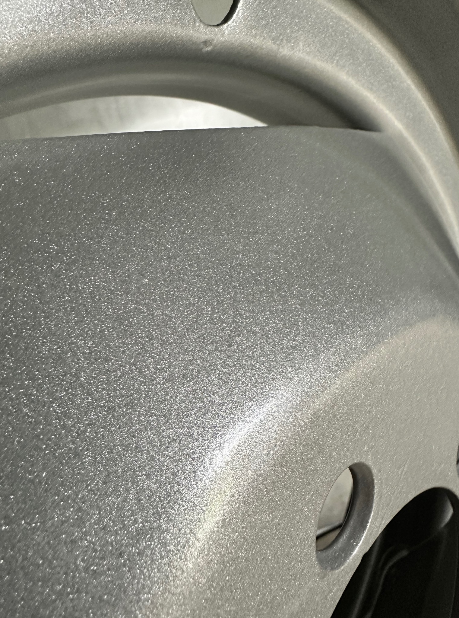 Szczegółowe ujęcie fragmentu felgi samochodowej po obróbce. Srebrzysta, metaliczna powierzchnia z widocznymi otworami montażowymi.