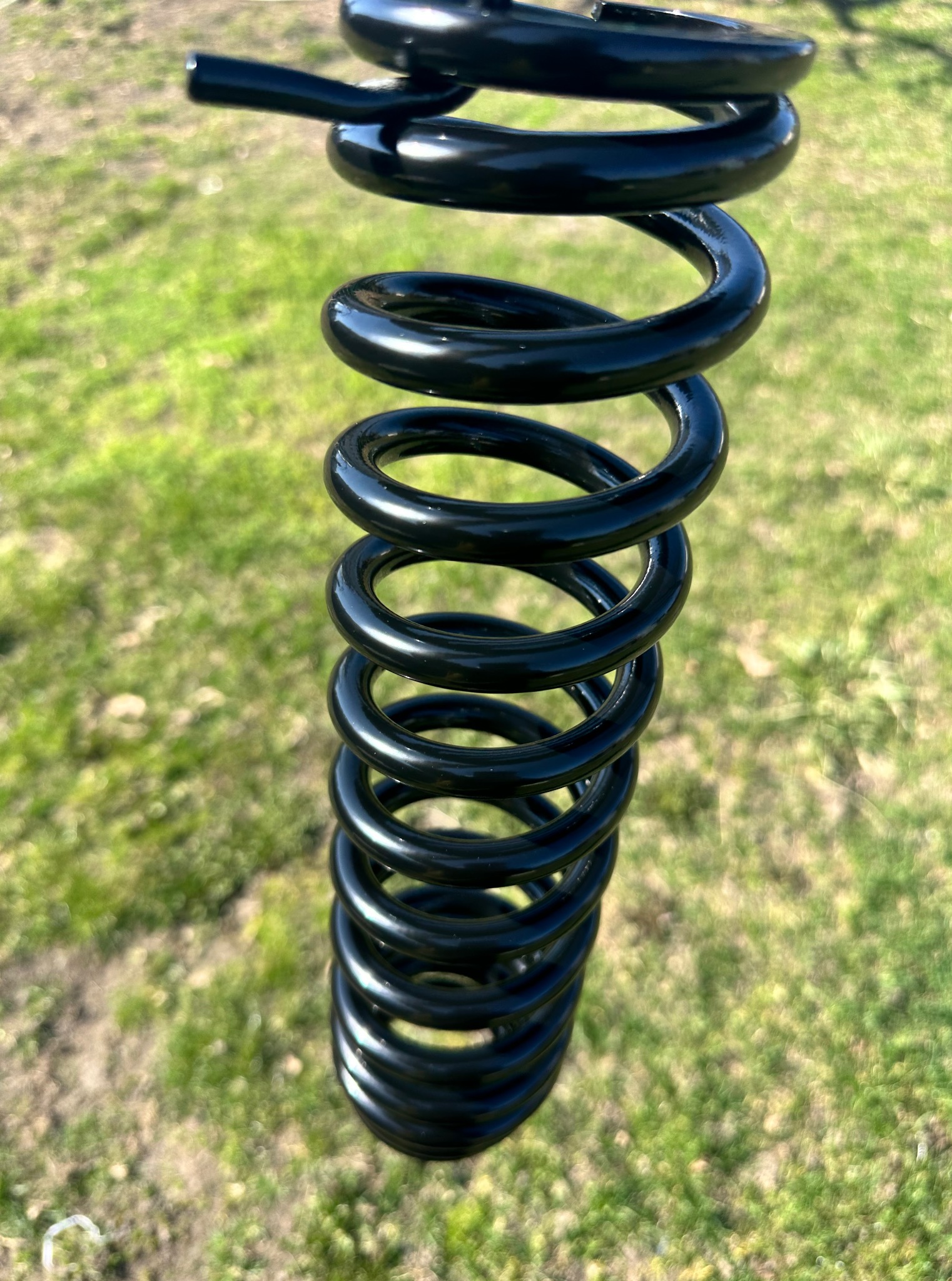 Błyszcząca, czarna, metalowa sprężyna na tle zielonej trawy. Metal precyzyjnie wykończony, gładki. Widoczny efekt finalny obróbki.