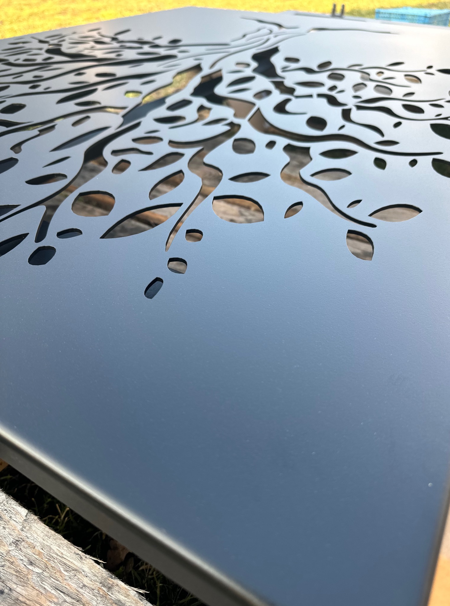 Metalowy panel dekoracyjny z wyciętym wzorem drzewa, pomalowany na gładki, ciemny kolor. Widoczne detale cięcia i faktura materiału.