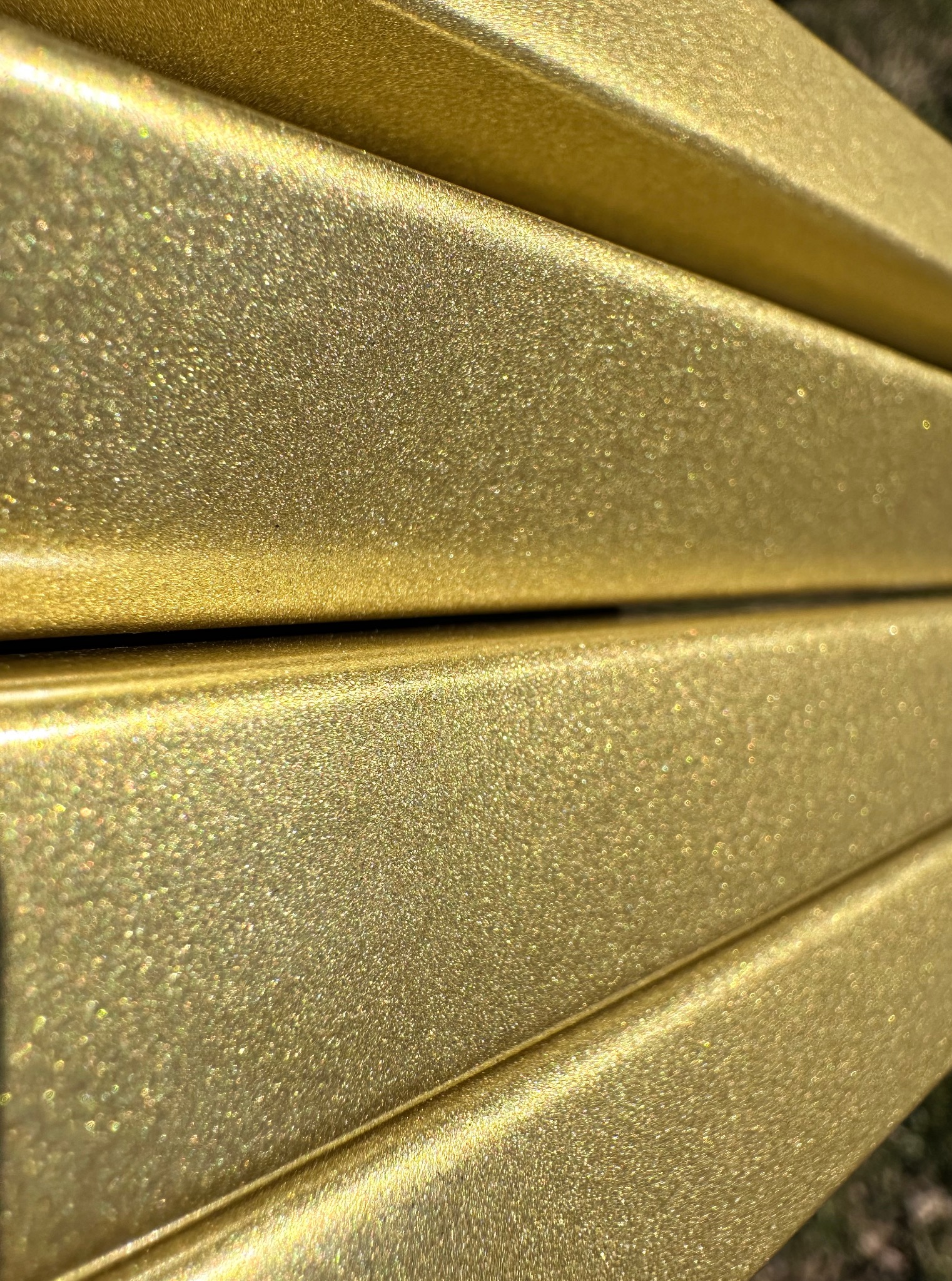 Błyszczące, złote profile pomalowane proszkowo, z efektem brokatu, ułożone równolegle. Widoczne delikatne odbicia światła i faktura powierzchni.