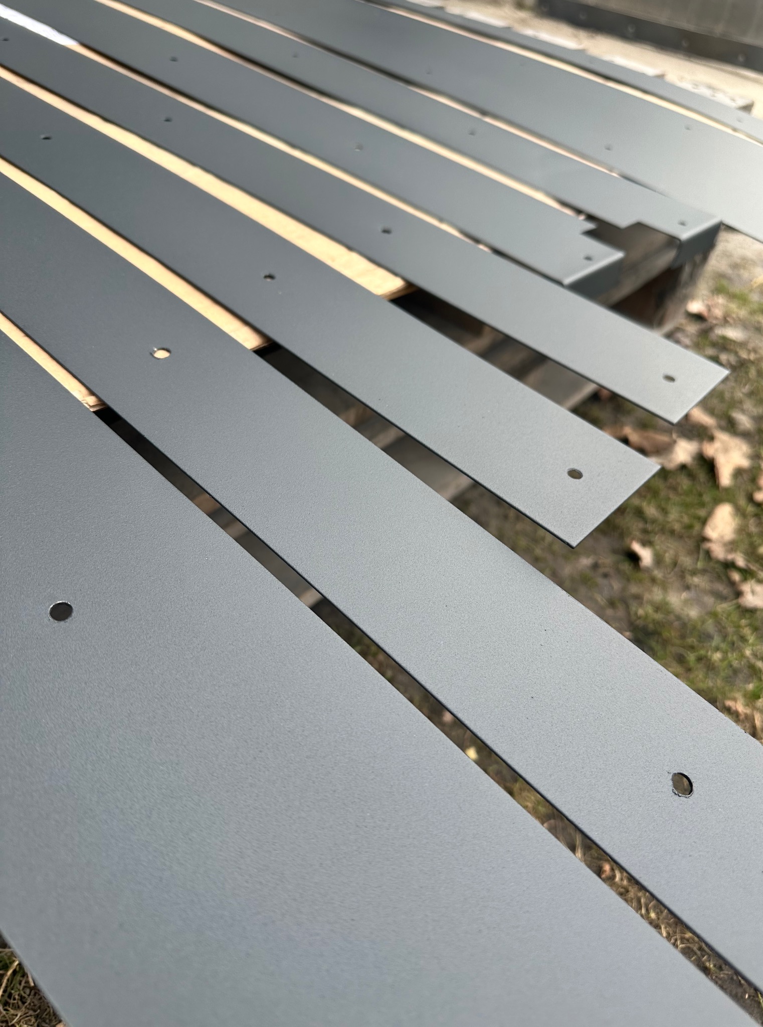 Szare, metalowe elementy z otworami, ułożone równolegle na drewnianej palecie na zewnątrz. Powierzchnia elementów jest jednolita, matowa i gładka.