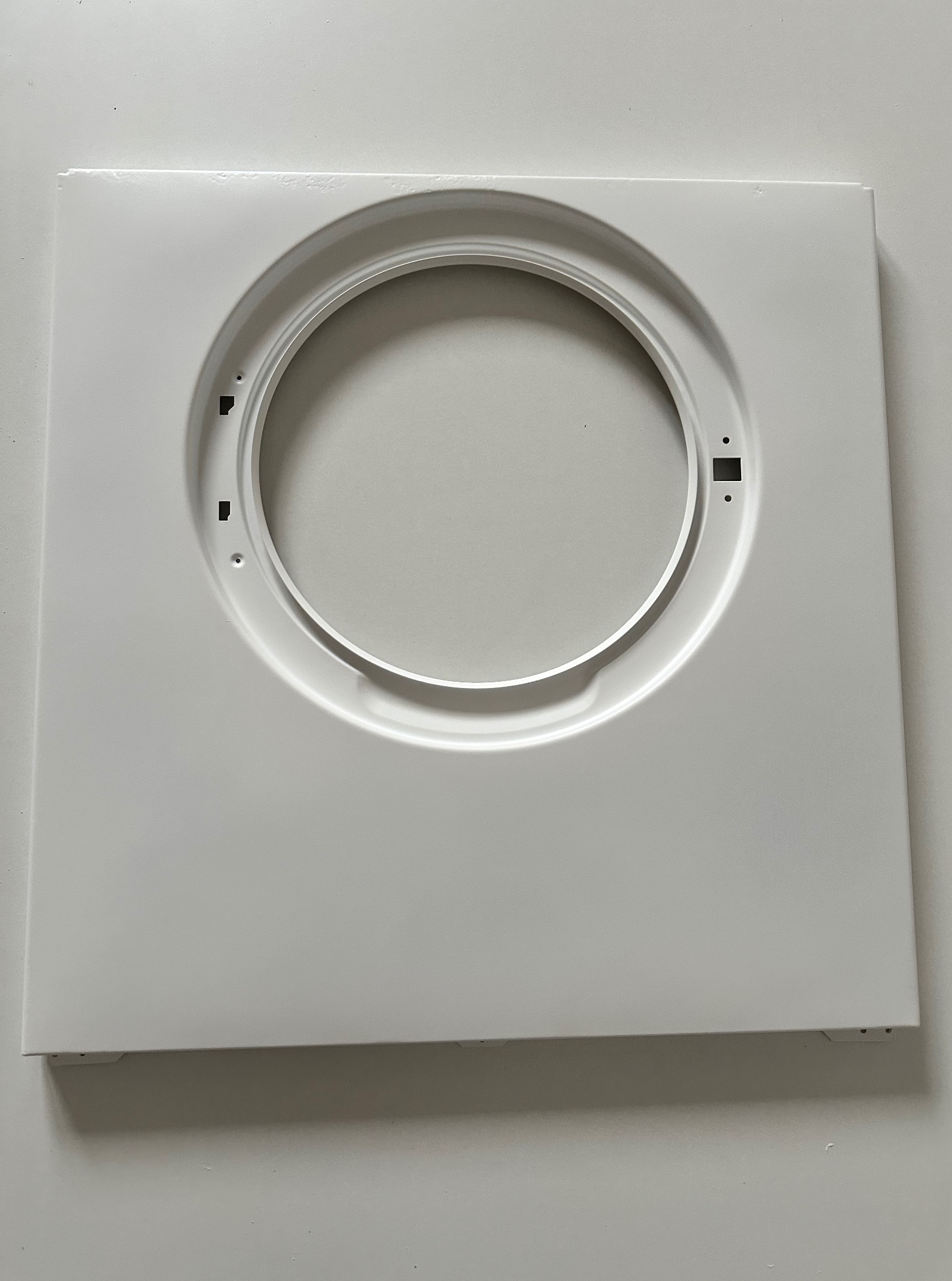 Biały, metalowy panel z okrągłym otworem pośrodku, leżący na jasnym tle. Widoczne drobne detale i otwory montażowe. Gładka powierzchnia.