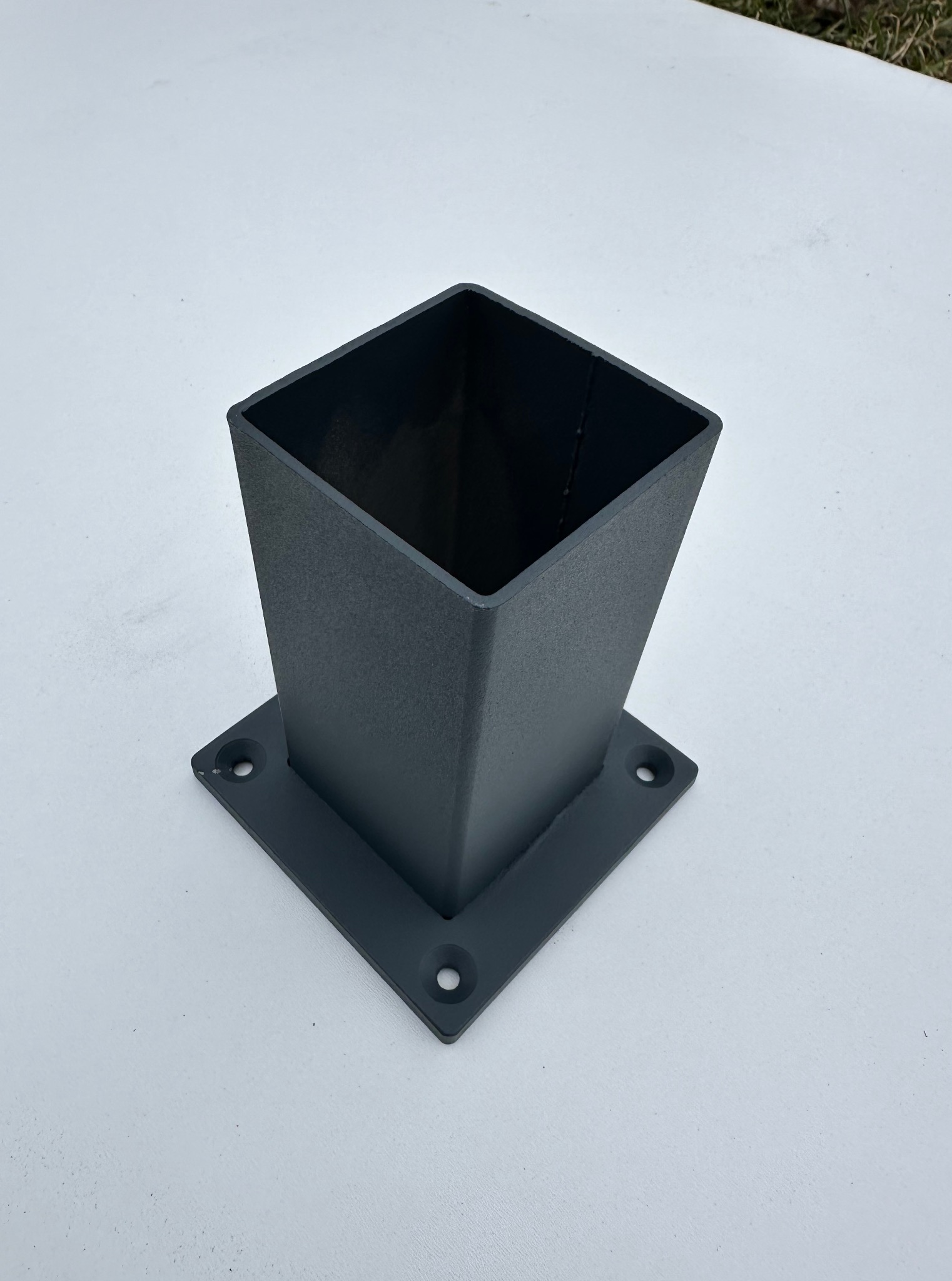 Metalowa podstawa słupka o kwadratowym przekroju, w kolorze grafitowym, z widocznymi otworami montażowymi, na jasnym tle. Detal konstrukcyjny.