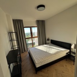 SunSet - Jasny pokój z parkietem w jodełkę: łóżko, regał i okno z widokiem na zieleń. Promienie słońca oświetlają materac, tworząc ciepłą atmosferę.