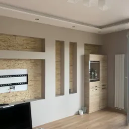 Wykończony salon z dekoracyjną ścianą z kamienia i wnękami, gotowy uchwyt na TV. Jasne ściany, drewniana podłoga i szafka. Widok na balkon.