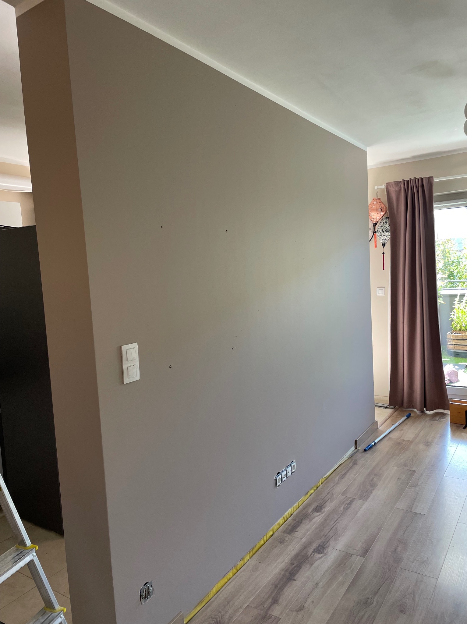Ściana w odcieniach szarości po malowaniu, z widocznymi gniazdkami elektrycznymi i listwą przypodłogową zabezpieczoną taśmą malarską. W tle fragment okna z zasłoną.