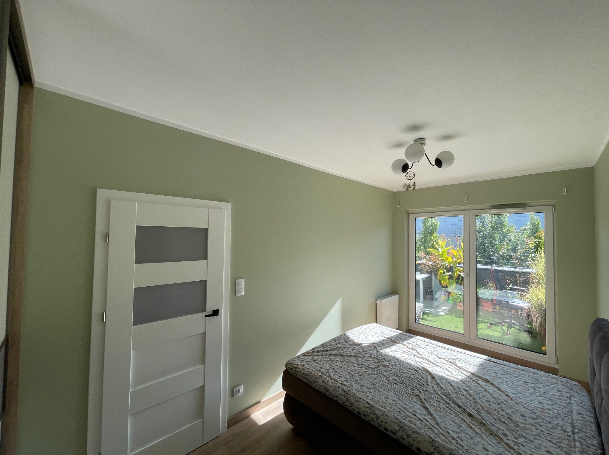 Jasnozielony pokój po malowaniu: ściany, białe drzwi z przeszkleniami, łóżko, okno z widokiem na ogród. Minimalistyczny wystrój.