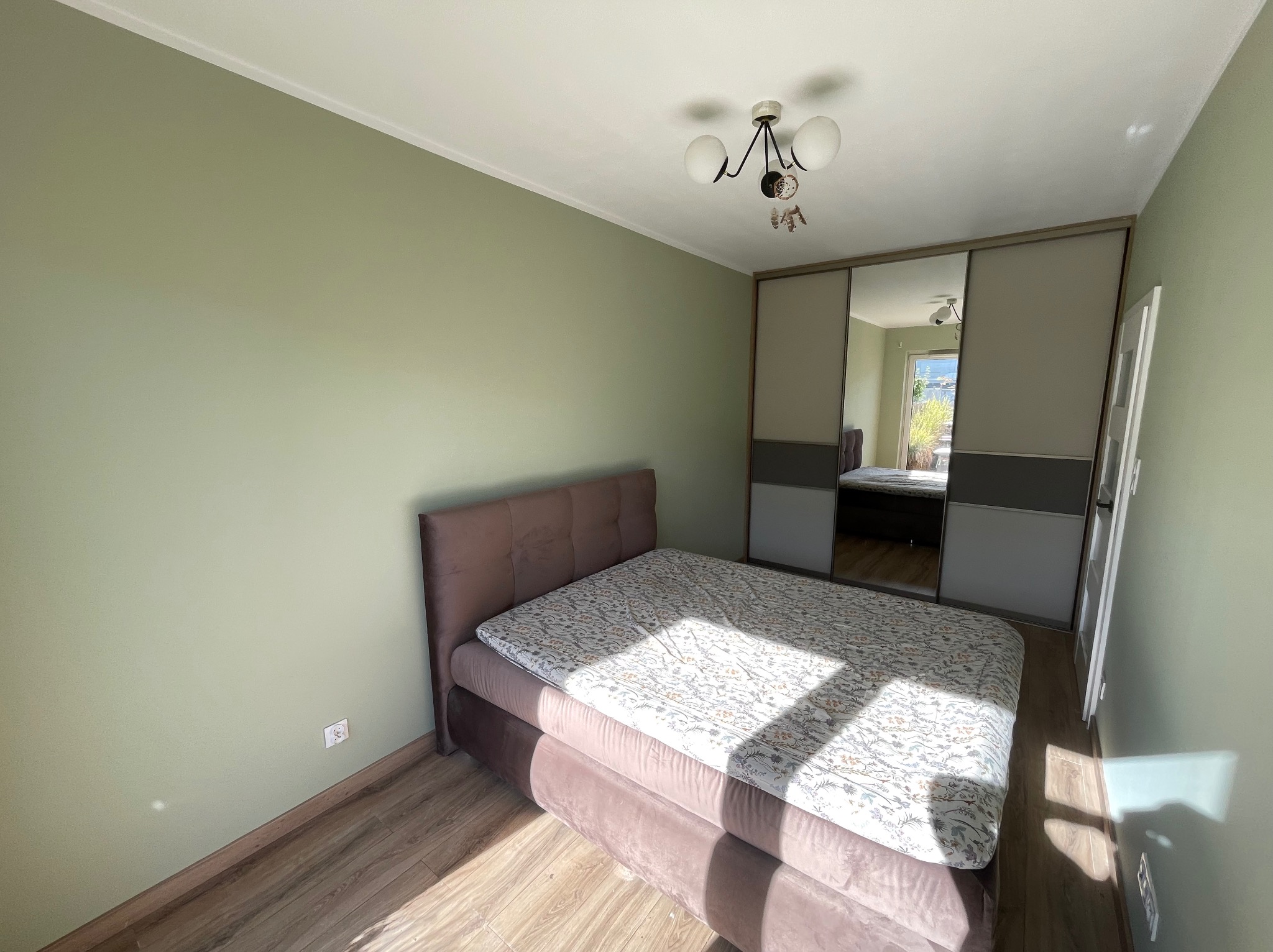 Jasna sypialnia po malowaniu: oliwkowa ściana, drewniana podłoga, łóżko z wezgłowiem w kolorze pudrowego różu i szafa z lustrzanymi drzwiami przesuwnymi.