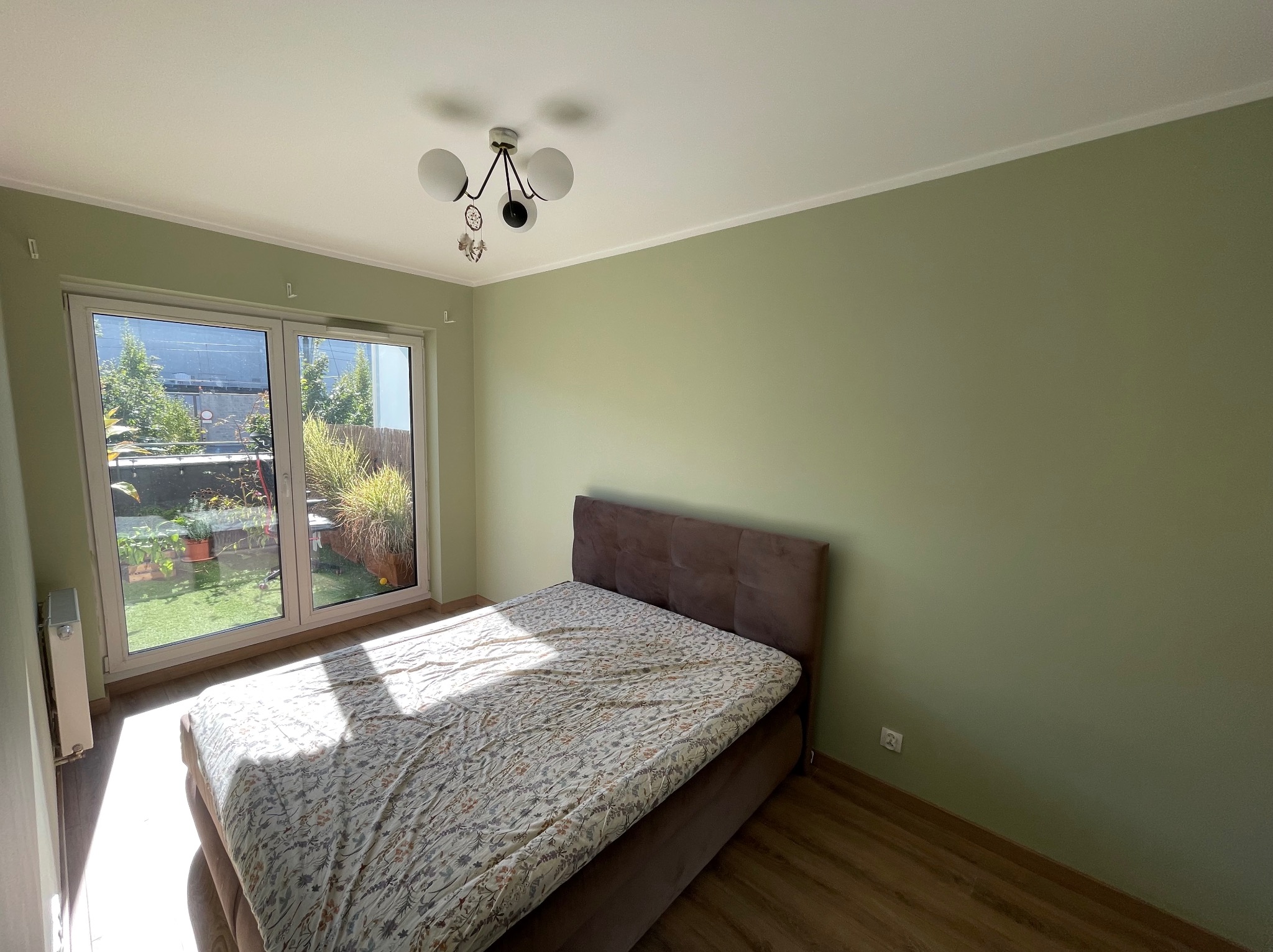 Pokój po malowaniu: zielone ściany, brązowe łóżko, widok na ogród przez okno balkonowe. Świeżo pomalowane ściany w odcieniu zieleni.