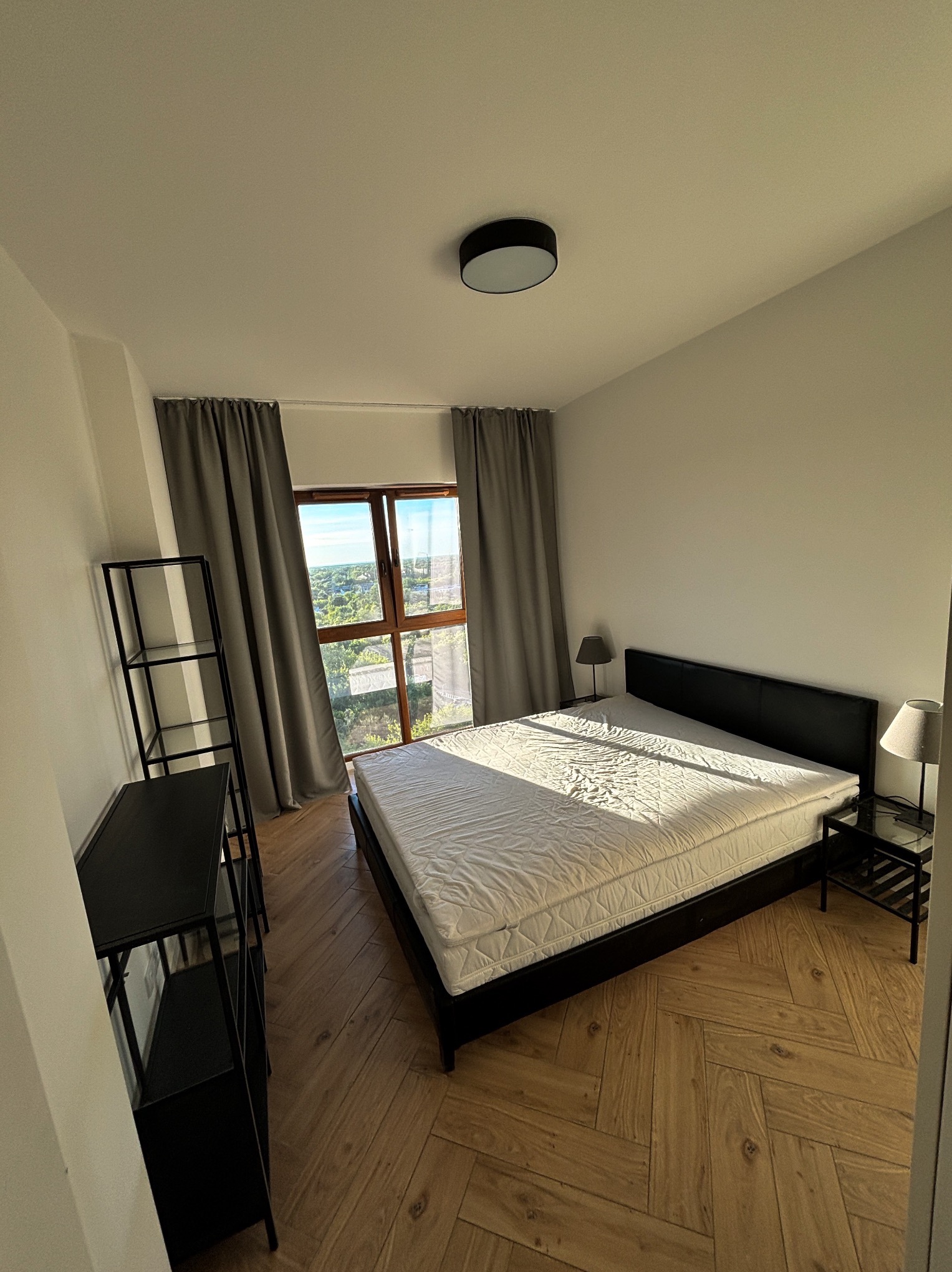 Jasny pokój z parkietem w jodełkę: łóżko, regał i okno z widokiem na zieleń. Promienie słońca oświetlają materac, tworząc ciepłą atmosferę.