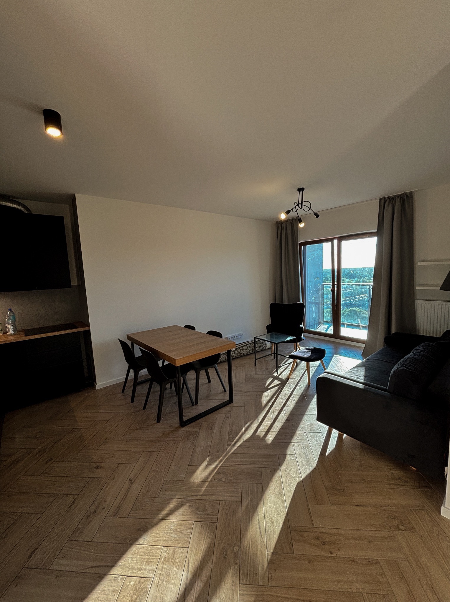 Jasne, przestronne wnętrze mieszkania z parkietem w jodełkę, czarnymi meblami i dużym oknem balkonowym z widokiem na zieleń. Promienie słońca padają na podłogę.