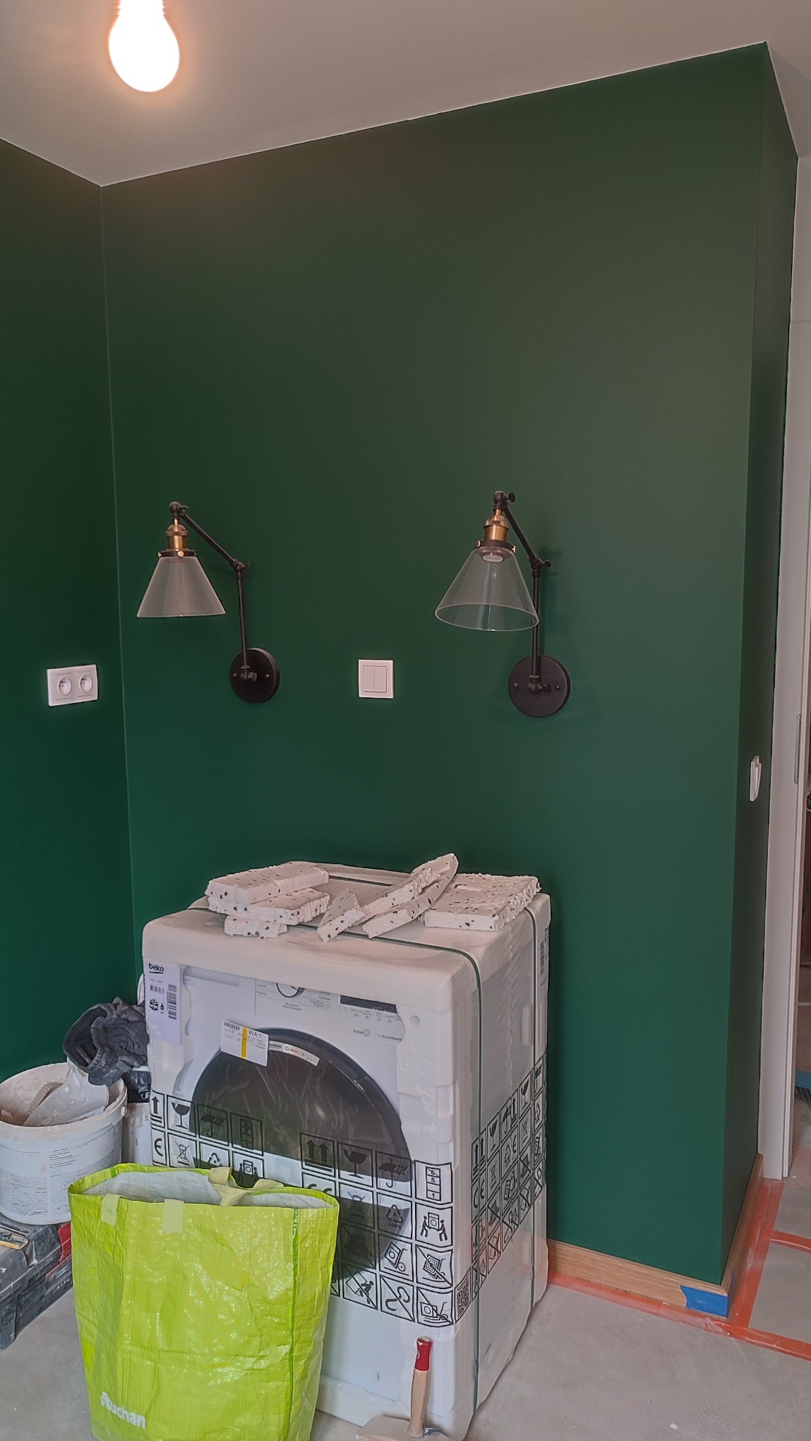 Ściana w głębokim odcieniu zieleni z dwoma kinkietami z kloszami, sprzęt AGD w folii zabezpieczającej i torba budowlana. Widoczne gniazdka i włącznik.