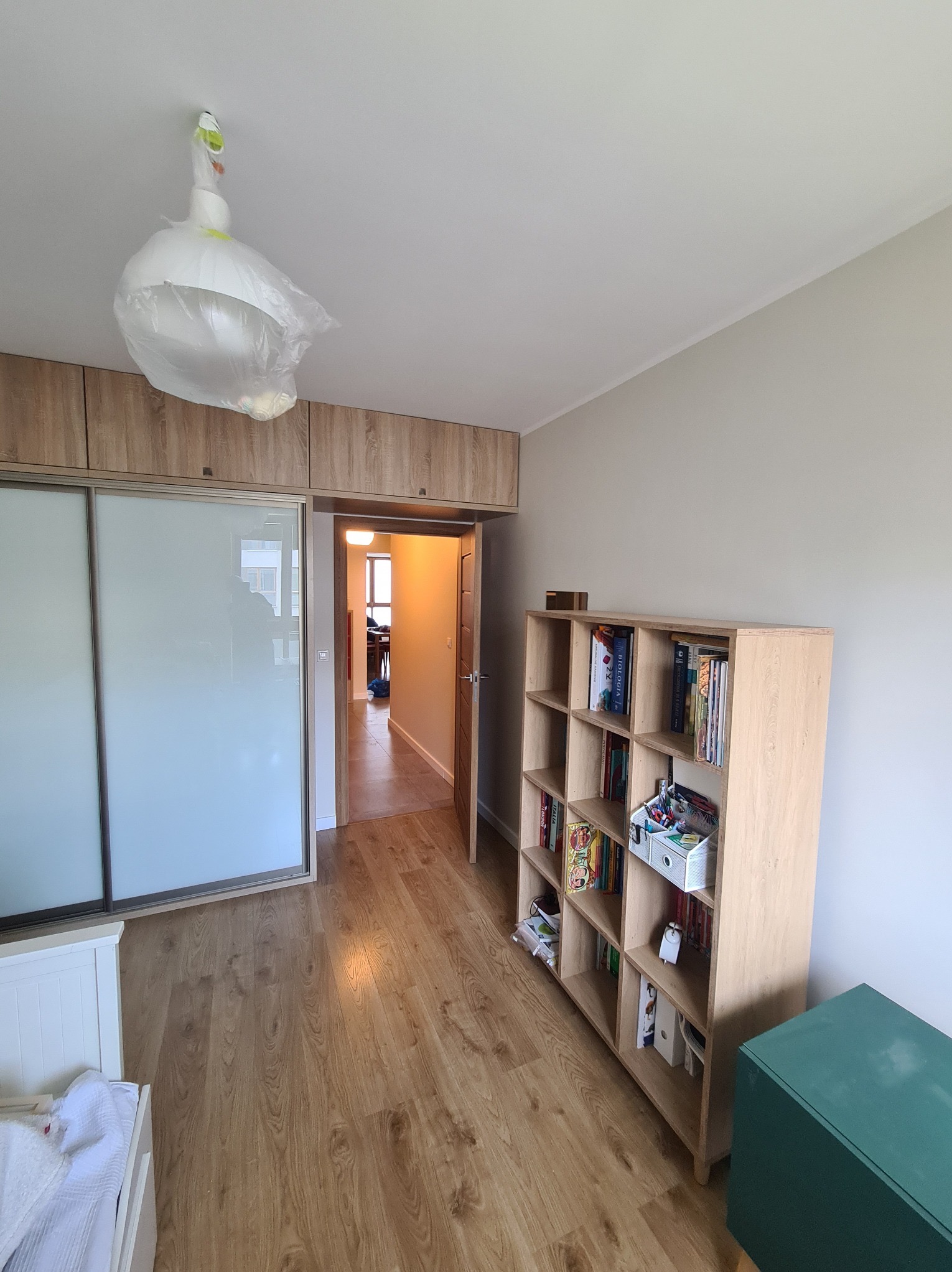Jasne wnętrze pokoju z drewnianą podłogą, regałem z książkami i szafą w zabudowie. Lampa zabezpieczona folią ochronną przed remontem. Widok na korytarz.