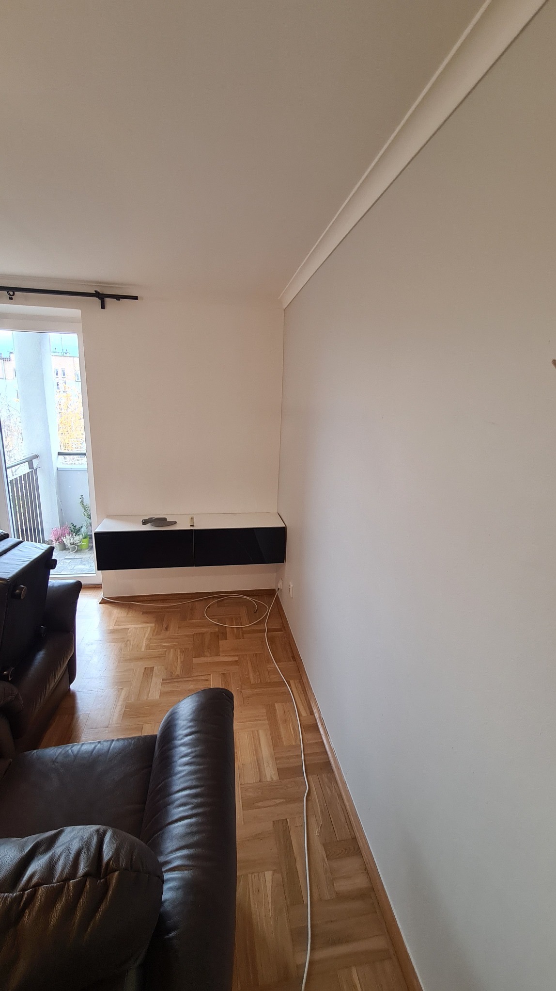 Jasne wnętrze po malowaniu ścian na neutralny odcień szarości, z widoczną listwą sufitową, parkietem i częściowo widocznym, skórzanym fotelem.