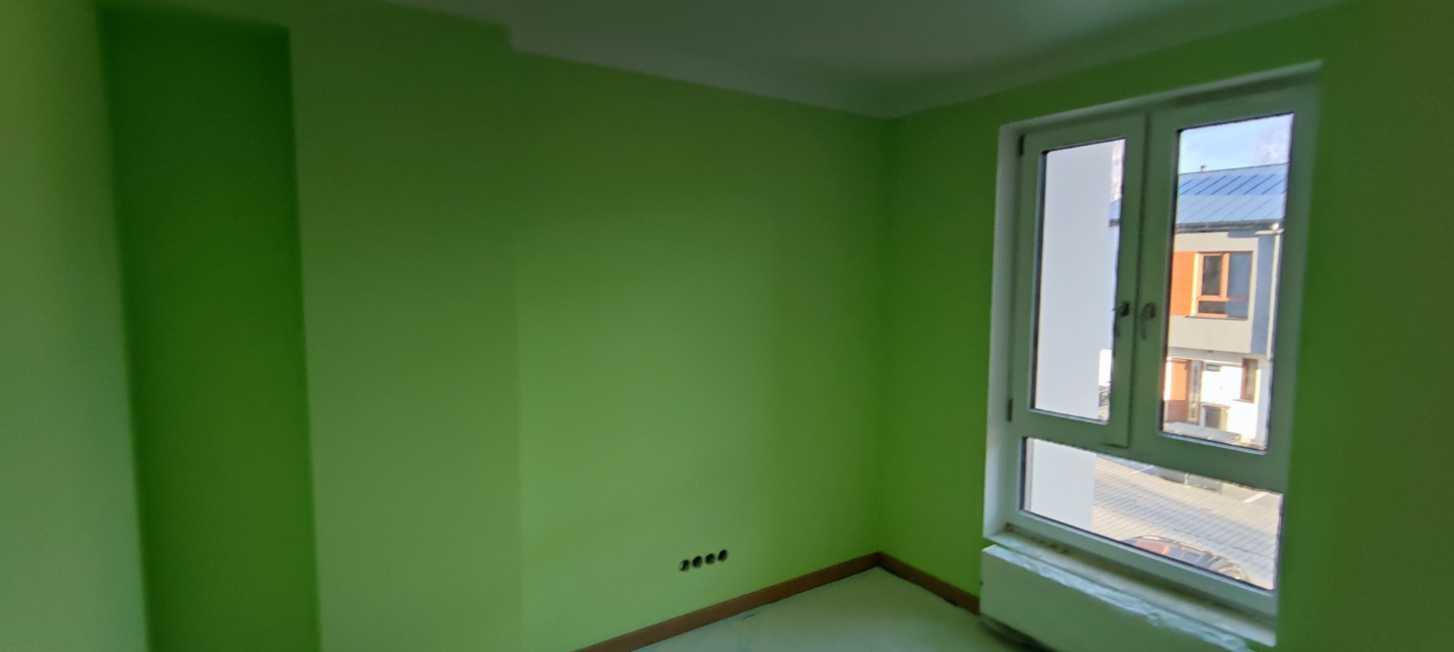 Pokój po malowaniu na zielono, widoczne gniazdka elektryczne i białe okno z widokiem na budynek. Świeżo pomalowane ściany, nowa kolorystyka wnętrza.