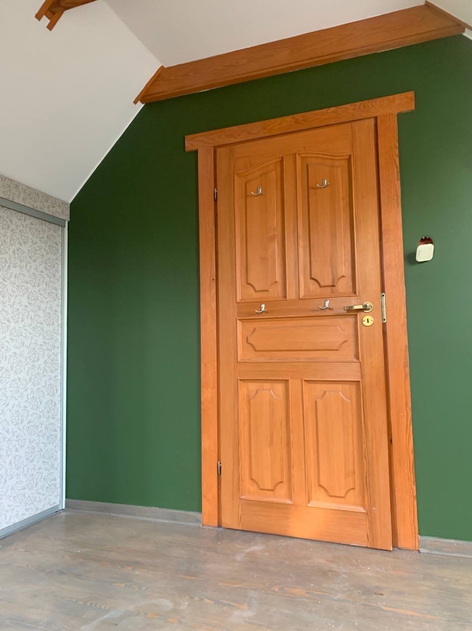 Drewniane drzwi z haczykami w zielonym pokoju z ukośnym sufitem i przesuwną szafą. Ściana pomalowana na jednolity, ciemnozielony kolor.