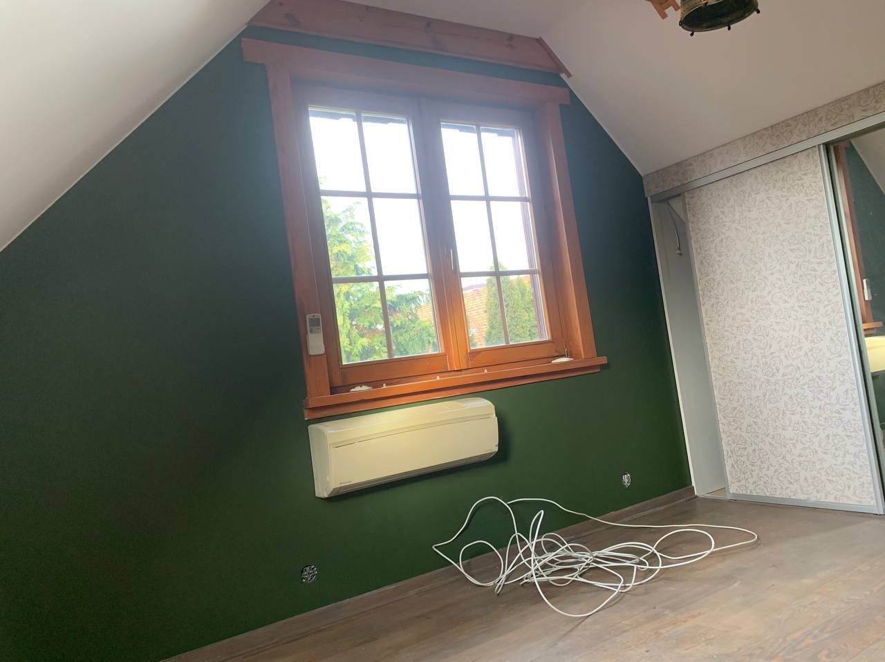 Pomieszczenie na poddaszu po malowaniu ścian na ciemnozielony kolor. Widoczne drewniane okno, klimatyzator i zwisające kable.
