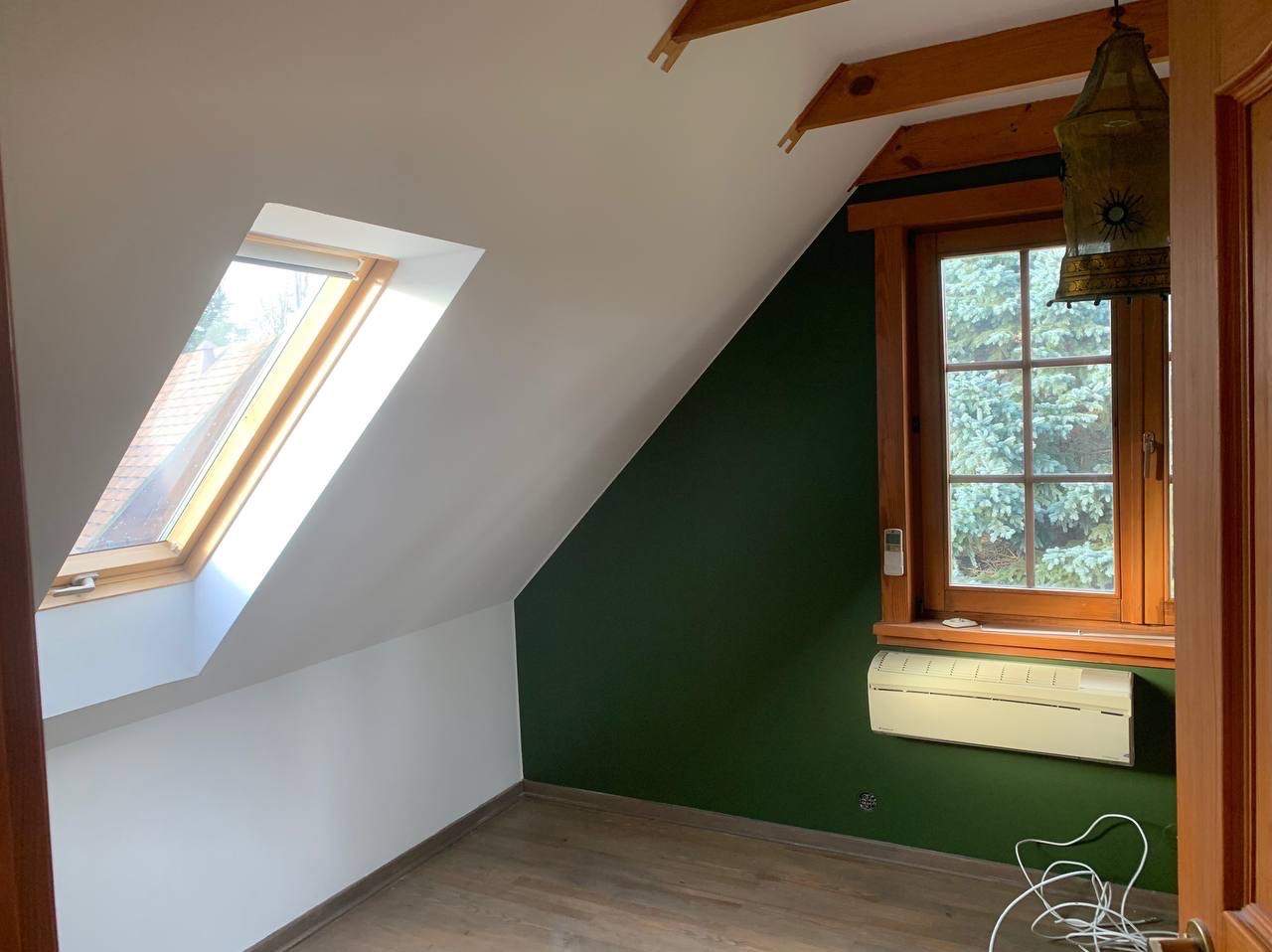 Pokój na poddaszu po malowaniu: białe skosy, ciemnozielona ściana, drewniane okna. Na ścianie klimatyzator, na suficie drewniane belki i ozdobna lampa.